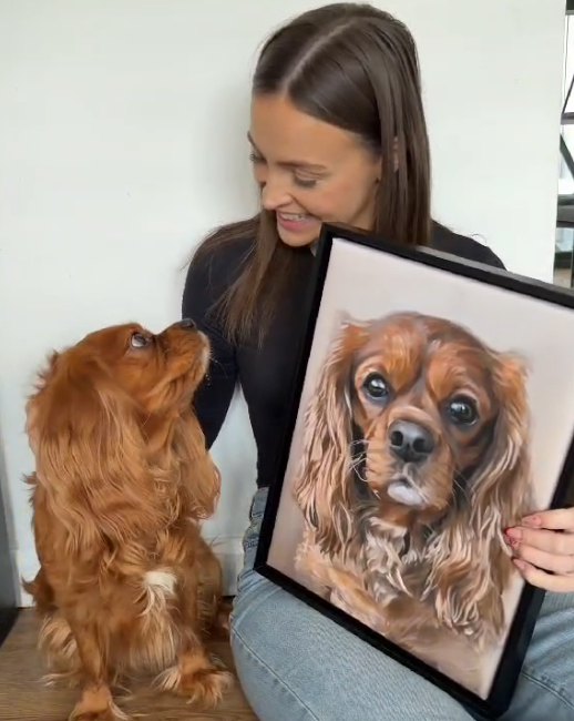 Woman holding a framed portrait of a dog next to a real dog.